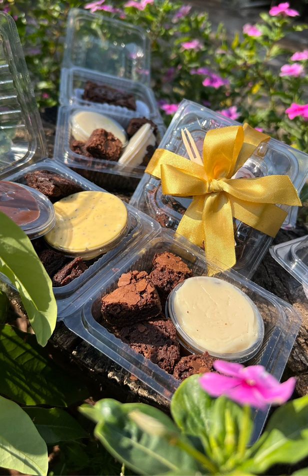 Potinhos de brownie com recheios variados e uma caixa com laço dourado, sobre um toco de madeira cercado de flores